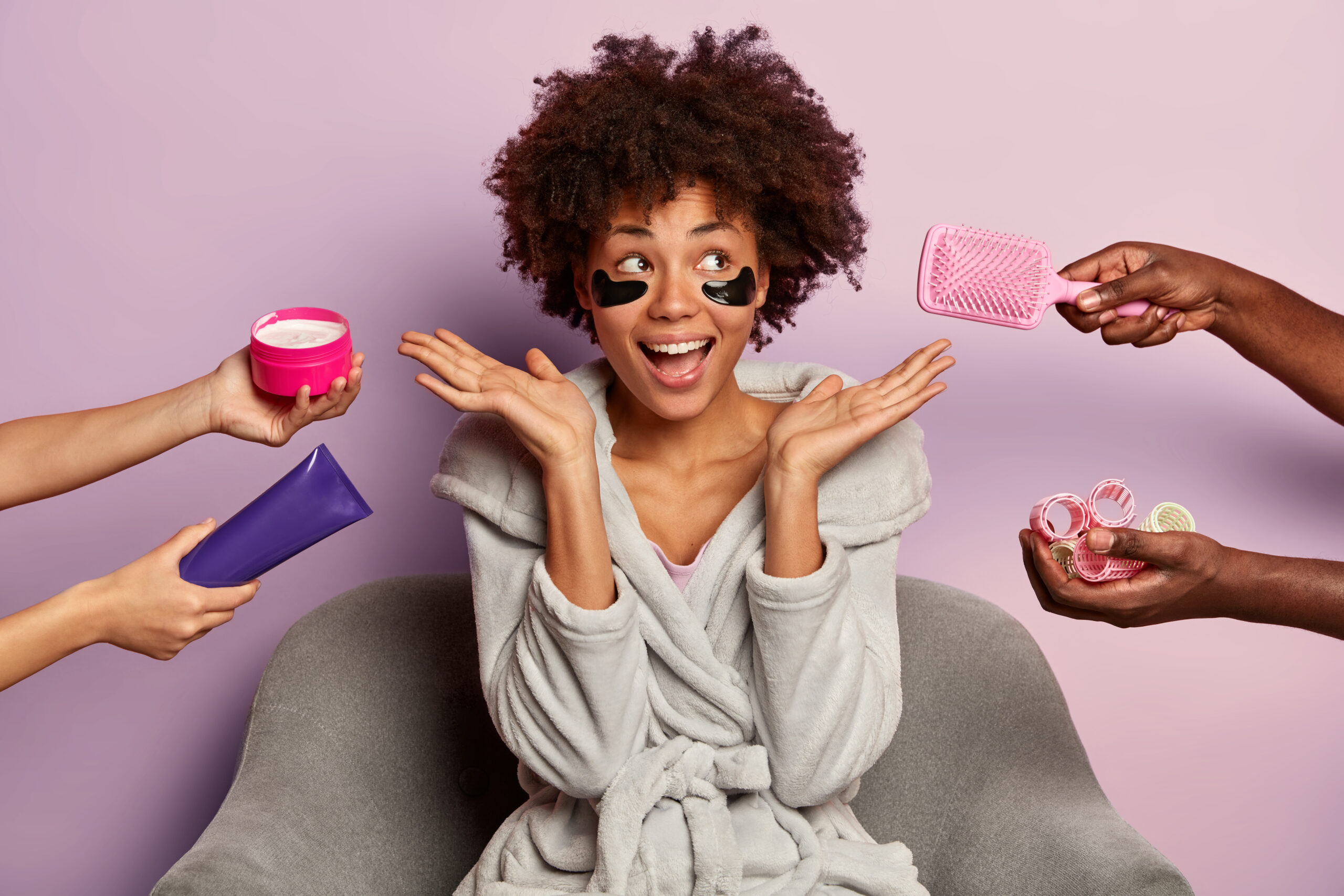 beauty spa woman with afro curly hairstyle, wears domestic clothes, poses on armchair, surrounded with humans hands holding cosmetic cream, haircomb and curlers, laughs from positive emotions